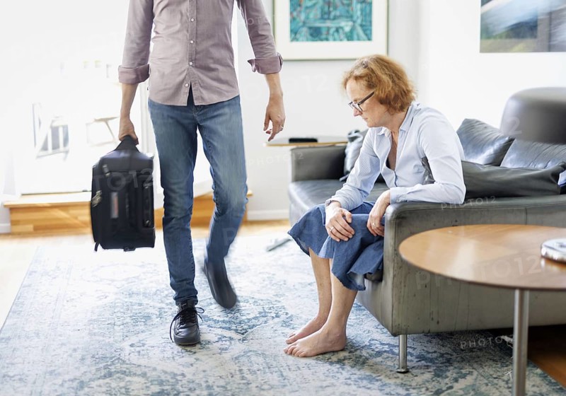 Symbolic photo on the subject of separation in a partnership. A man carries a suitcase through a living room while his woman sits depressed on the sofa. Berlin, 13.08.2024