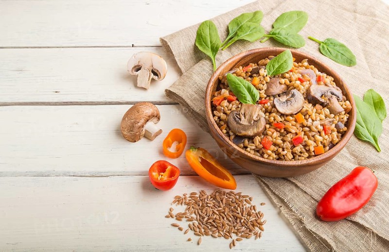 Spelt (dinkel wheat) porridge with vegetables and mushrooms in wooden bowl on a white wooden background and linen textile. Side view, copy space. Russian traditional cuisine
