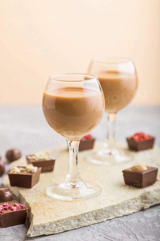 Sweet chocolate liqueur in glass with chocolate candies on a gray concrete background and stone slate board. Side view, close up, selective focus
