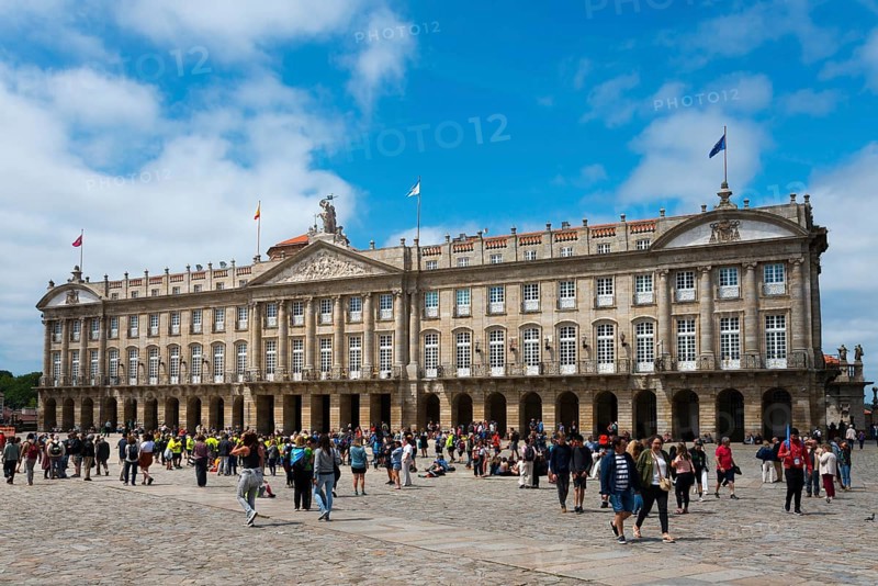 A large baroque stone structure with many windows and visitors in front of it in an open square under a blue sky, Pazo de Raxoi, Concello de Santiago de Compostela, palace, town hall, Praza do Obradoiro, town square, Santiago de Compostela, autonomous community of Galicia, place of pilgrimage, destination of the Way of St James, Spain, Europe