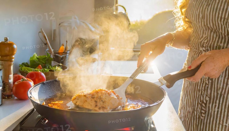 A person is cooking chicken in a pan on a stove. The pan is filled with a red sauce and the chicken is surrounded by steam, AI generated