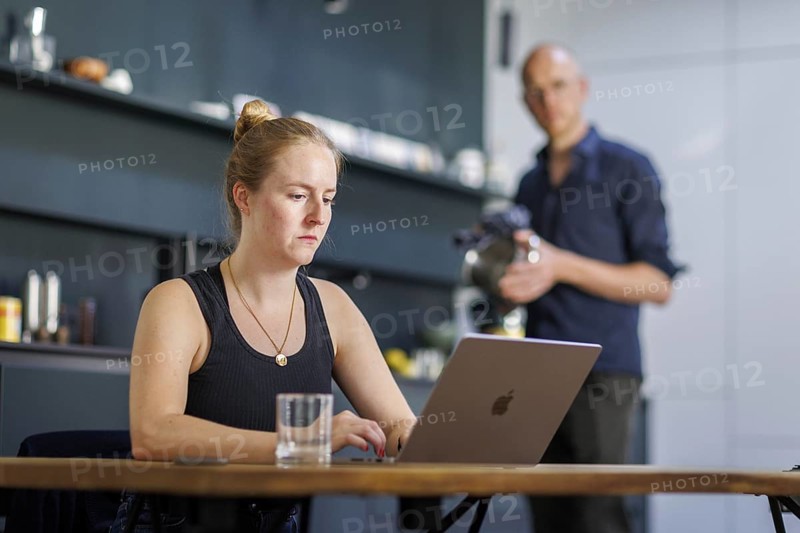 Symbolic photo on the subject of division of labour for couples in the household. A woman sits at a laptop in a kitchen while a man washes dishes in the background. Berlin, 13.08.2024
