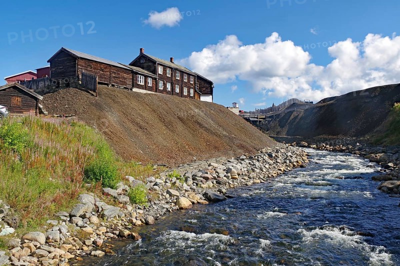 A rushing river next to old buildings on a hill under a sky with white clouds, old mine, overburden, copper ore, Unesco World Heritage Site, mining town, Röros, Norway, Europe