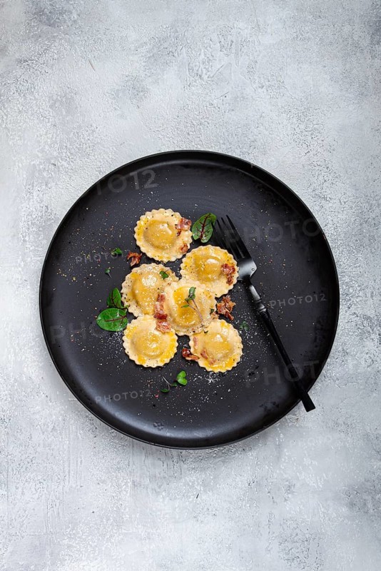 Fresh ravioli, with butter, spices and herbs, fried bacon, on a black plate, top view, dark background, no people