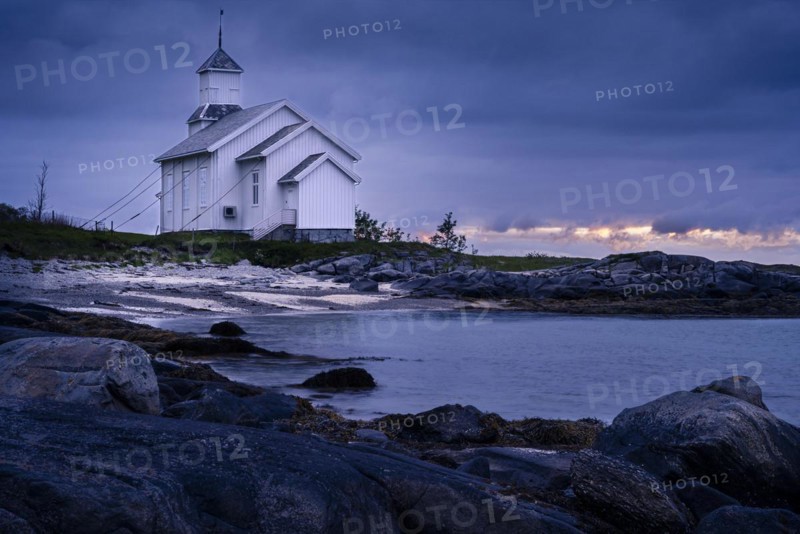 The white church of Gimsoy (Gimsoysand church) by the sea, rocks in the foreground. At night at the time of the midnight sun. Cloudy sky, a narrow strip of light in the sky. Early summer. Gimsoya, Lofoten, Norway, Europe