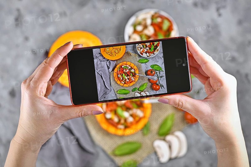 Taking photo of food with hands holding mobile phone with picture of vegan baked Red kuri squash vegetable filled with bell pepper, tomatoes and mushrooms surrounded by ingredients