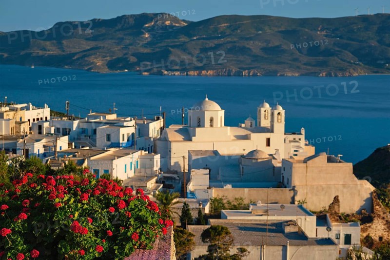 View of Plaka village on Milos island over red geranium flowers on sunset. Plaka town, Milos island, Greece. Focus on flowers