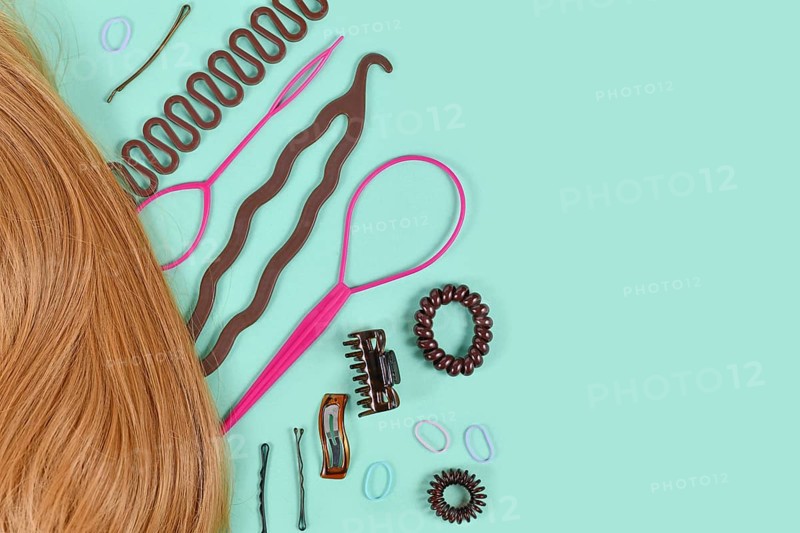 Hair styling tools like bun maker, braid tool, ponytail style maker, hair clip, rubber bands and pins next to blond hair with copy space