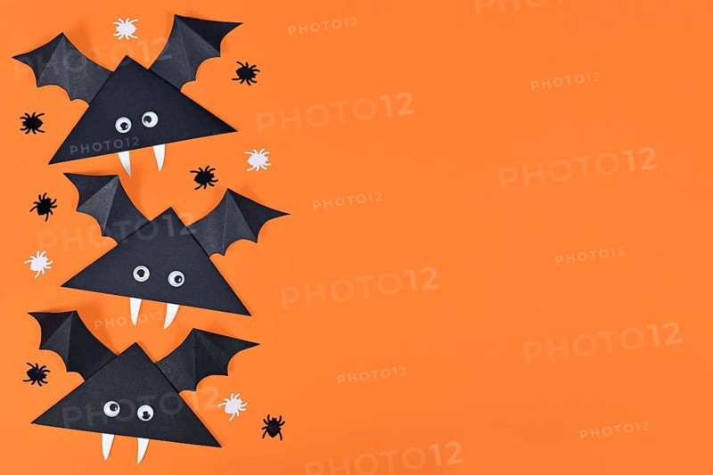 Cute paper vampire bats with funny googly eyes on side of orange Halloween background with empty copy space