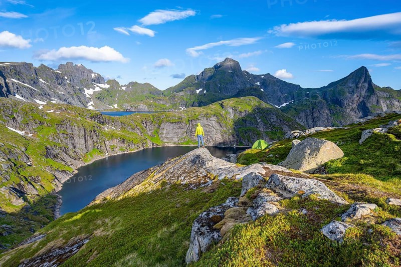 Mountaineer standing on a rock with green camping tent, mountain landscape with lake Tennesvatnet, at sunrise, behind peak of Hermannsdalstinden, Moskenesøya, Lofoten, Nordland, Norway, Europe