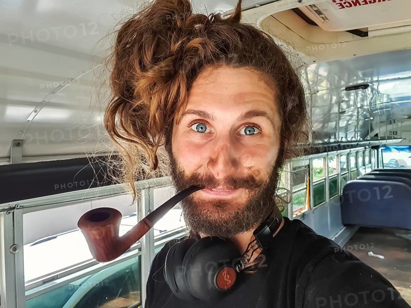 Close up portrait shot of a curious looking young man with long hair, big headphones and a wooden vintage pipe inside a bus being renovated