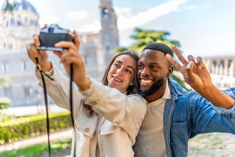 Multi-ethnic couple gesturing success while taking selfie with digital mirrorless camera in a park