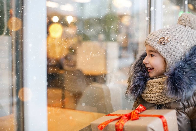 Portrait of joyful girl with a gift box for Christmas near the glass shop window in winter with snow on festive market with decorations and lights. Warm clothes, knitted hat, scarf and fur. Copy space