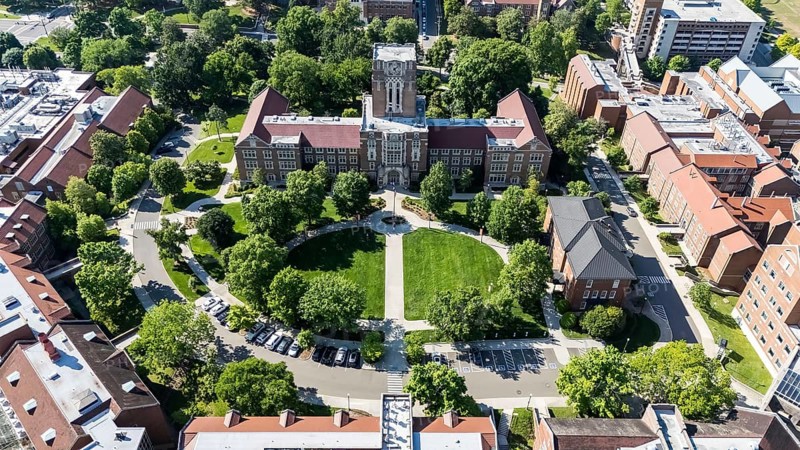 An aerial view of the University of Tennessee, Knoxville showcases a sprawling campus with lush greenery, historic buildings, modern research facilities, and athletic fields, nestled against the backdrop of the Tennessee River and Smoky Mountains