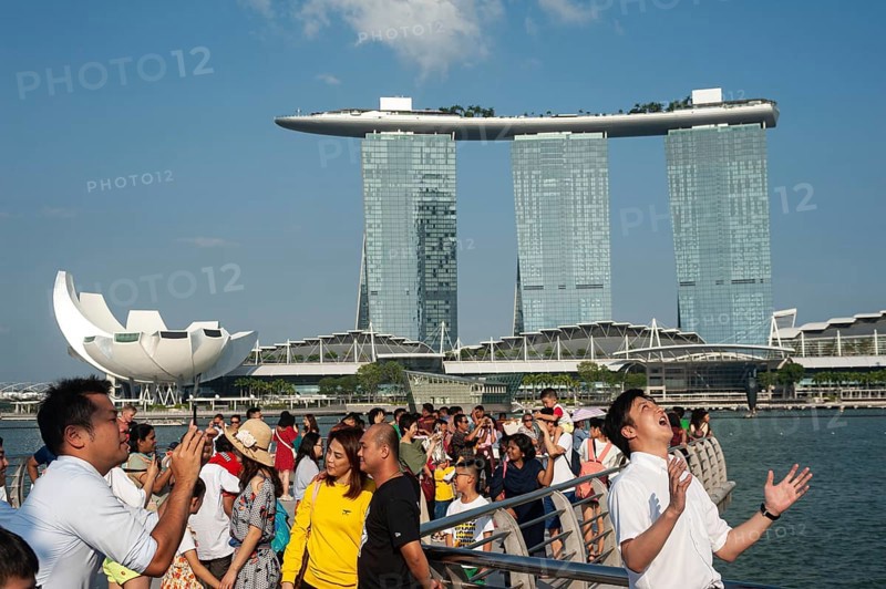 01.08.2019, Singapore, Republic of Singapore, Asia, Tourists pose for photos in Merlion Park on the banks of the Singapore River with the Marina Bay Sands Hotel in the background. The open mouth is meant to catch the stream of water from the Merlion figure, which is not in the picture, Asia