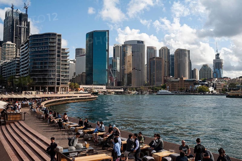 2019-09-25, Sydney, New South Wales, Australia, A view of the skyline of the business district around Circular Quay and the harbour. In the foreground, people sit in the Opera Bar along the waterfront, Oceania