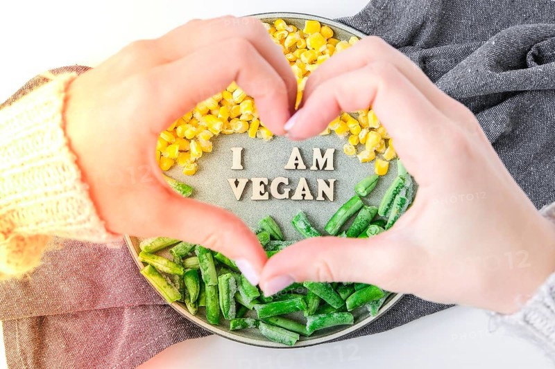 Female hands in shape of heart love I AM VEGAN text in plate. Veganism, vegetarian healthy lifestyle green beans yellow corn. Healthy eating vegan. Wooden letters