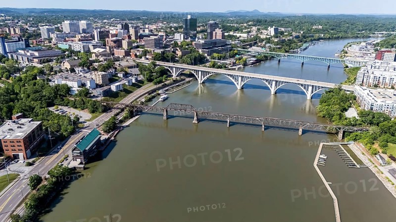An aerial view of Knoxville, Tennessee reveals a vibrant cityscape with a mix of historic and modern buildings, the Tennessee River weaving through downtown, lush green parks, and the distant Smoky Mountains framing the horizon