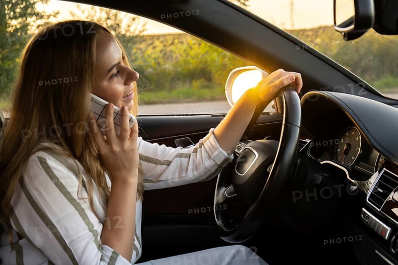 Happy young woman speaking by mobile phone while driving car. Business woman talking phone call in automobile. Unsafely risky driving. Concept of multitasking