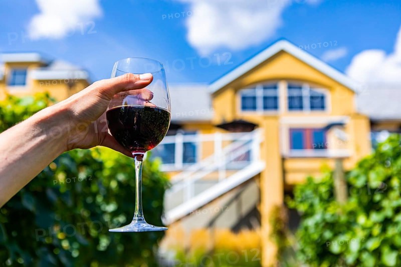 Man hand holding a glass of red wine selective focus close up view against big yellow wine tasting winery farm house background, blurred copy space