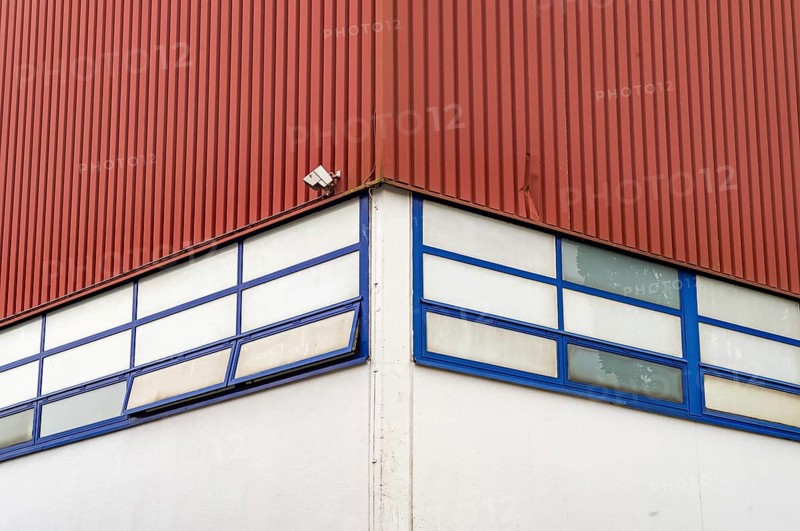 A single surveillance camera is mounted on the corner of a red industrial building with blue-framed windows
