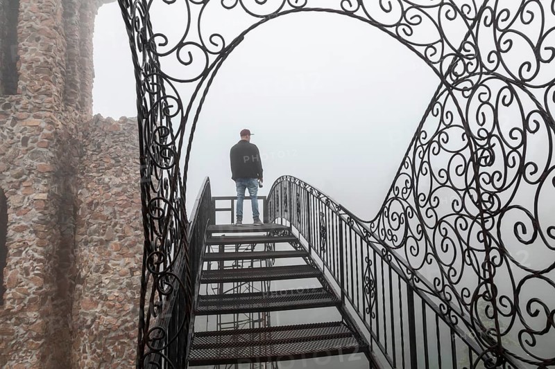 Rye, Colorado, A main stands on an open stairway, high above the ground, at Bishop Castle on a foggy morning. The Castle is an elaborate stone and metal structure and roadside attraction. It is being built by Jim Bishop in the mountains of San Isabel National Forest