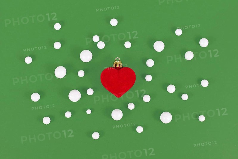 Red heart shaped Christmas tree bauble surrounded by snowballs on green background
