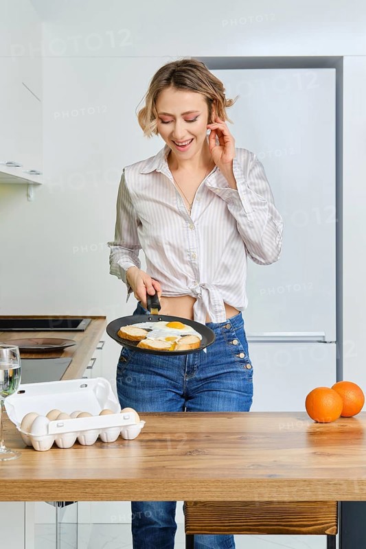 Happy young woman holds skillet with fried eggs and toasts in hand