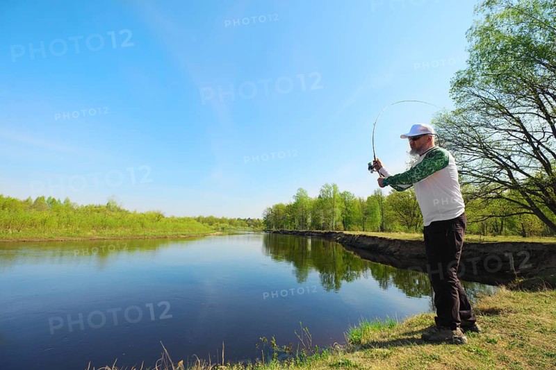 Fisherman with a spinning rod catching fish on a river at sunny summer day with green trees at background. Outdoor weekend activity. Photo with shallow depth of field taken at wide open aperture