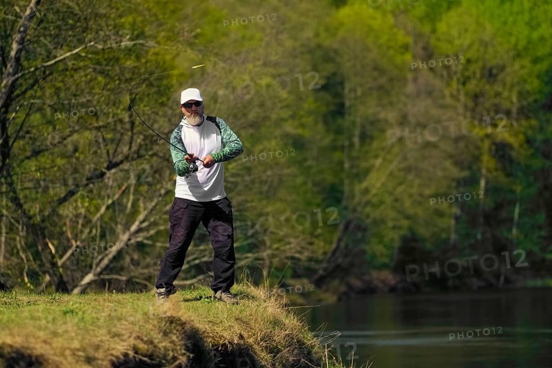 Fisherman throwing the bait into river standing on river bank in sunny day with green forest in background