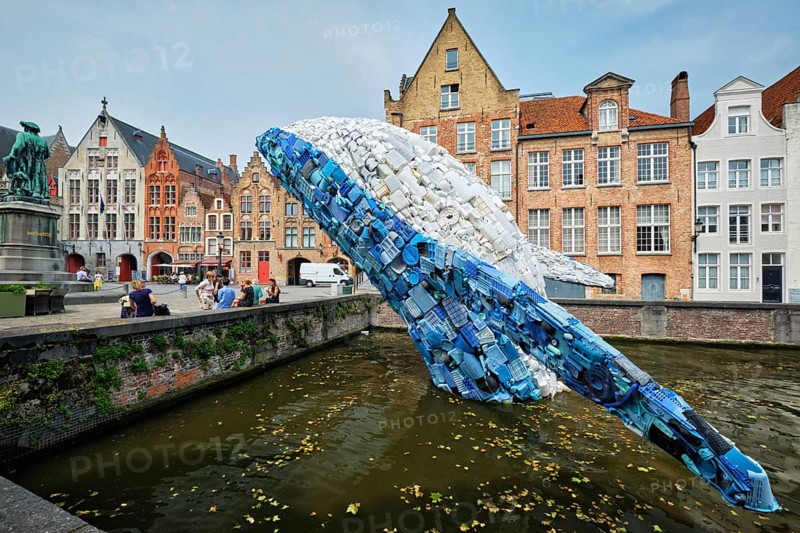 BRUGES, BELGIUM, MAY 29, 2018: Skyscraper or The Bruges Whale, whale rising up from the Canal installation made up of 5 tons of plastic waste pulled out of the ocean in front of Jan van Eyck statue