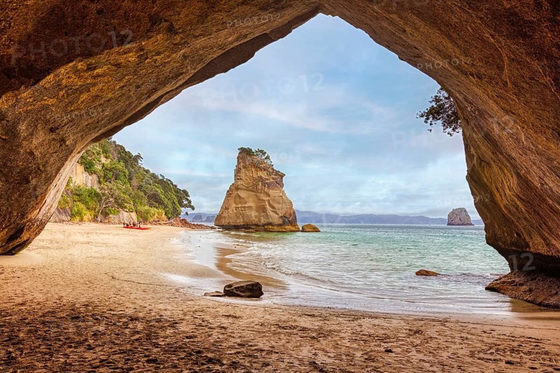 View from a cave opening onto a sandy beach and a solitary rocky outcrop in the sea, Cathedral Grove, Coromandel, New Zealand, Oceania