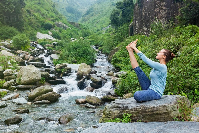 Yoga exercise outdoors, woman doing Ashtanga Vinyasa Yoga balance asana Ubhaya padangusthasana Big Double Toe Yoga Pose at tropical waterfall in Himalayas in India