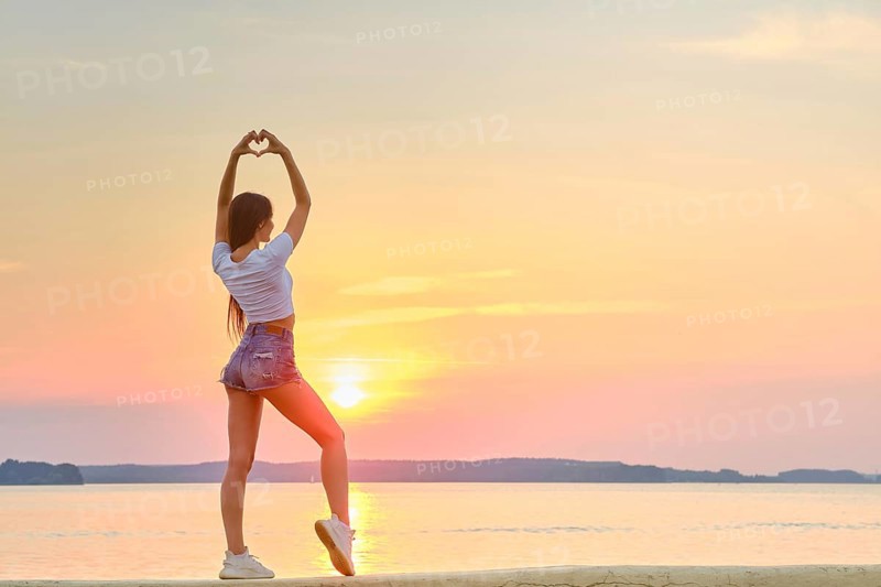 Woman raised her hands up and folded her palms in shape of heart against backdrop of sunset