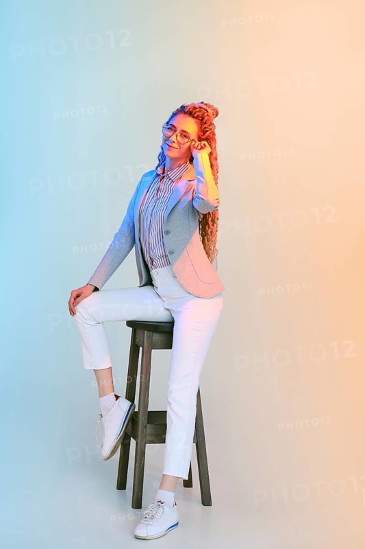 Full length portrait of young woman in jeans and jacket holding the bow of the glasses under colorful lights