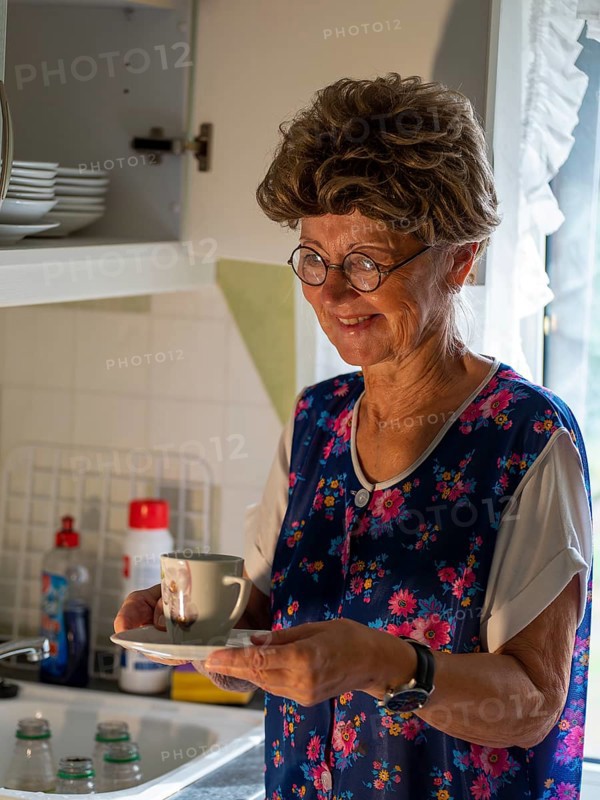 Granny with old smock apron, glasses and wig at the kitchen window in the kitchen, Germany, Europe