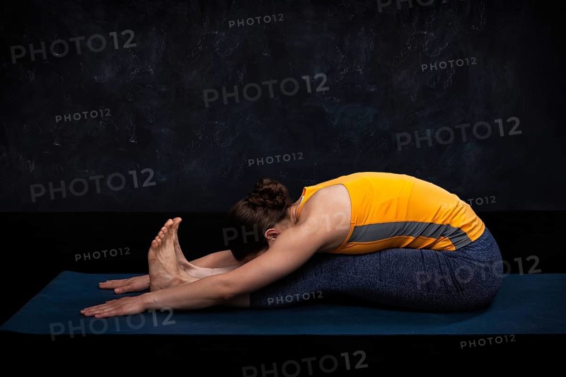 Woman doing Ashtanga Vinyasa Yoga back bending asana Paschimottanasana, seated forward bend on dark grunge background