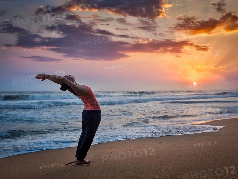 Young sporty fit woman doing yoga Sun salutation Surya Namaskar pose Hasta Uttanasana on tropical beach on sunset