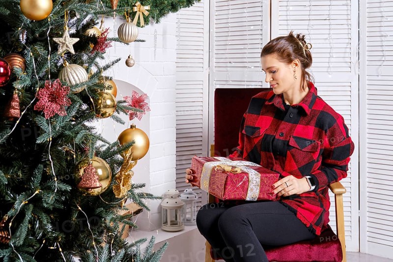 Young woman in warm plaid shirt and tight trousers looks at christmas gift box in hand