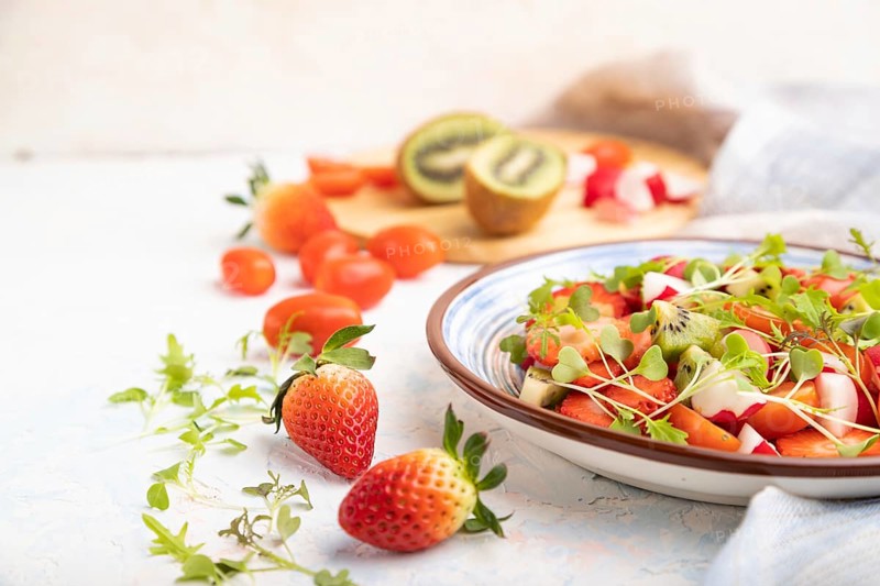Vegetarian fruits and vegetables salad of strawberry, kiwi, tomatoes, microgreen sprouts on white concrete background and linen textile. Side view, close up, selective focus