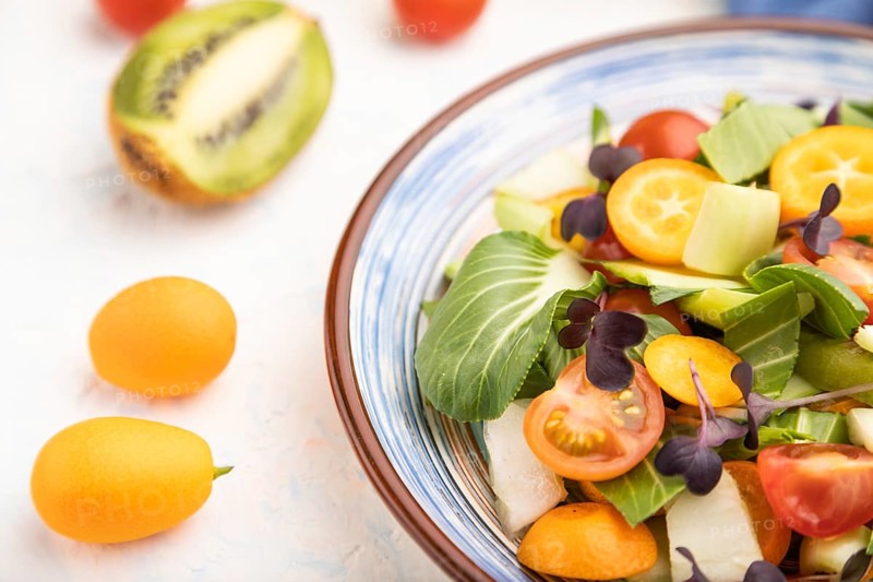 Vegetarian salad of pac choi cabbage, kiwi, tomatoes, kumquat, microgreen sprouts on a white concrete background and blue linen textile. Side view, close up, selective focus