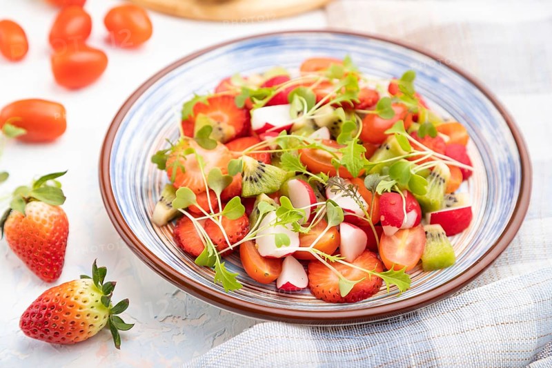 Vegetarian fruits and vegetables salad of strawberry, kiwi, tomatoes, microgreen sprouts on white concrete background and linen textile. Side view, close up, selective focus