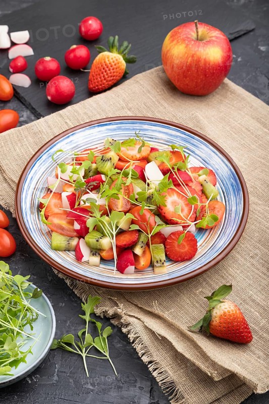 Vegetarian fruits and vegetables salad of strawberry, kiwi, tomatoes, microgreen sprouts on black concrete background and linen textile. Side view, close up