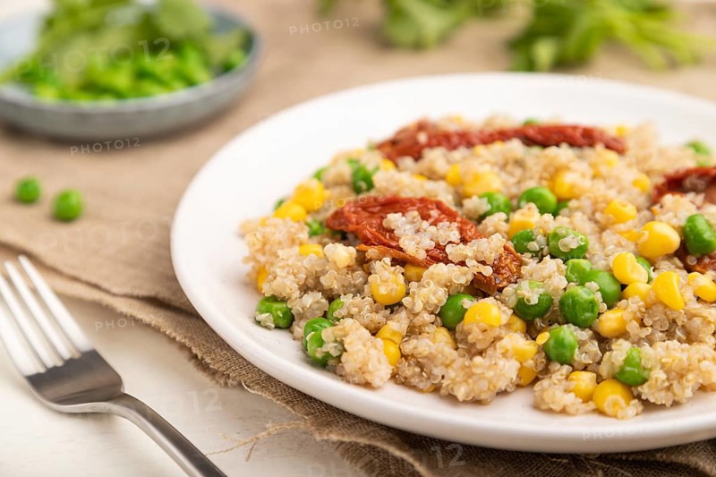 Quinoa porridge with green pea, corn and dried tomatoes on ceramic plate on a white wooden background and linen textile. Side view, close up, selective focus