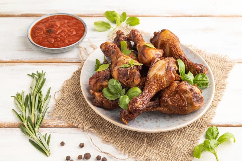 Smoked chicken legs with herbs and spices on a ceramic plate with linen textile on a white wooden background. Side view, close up