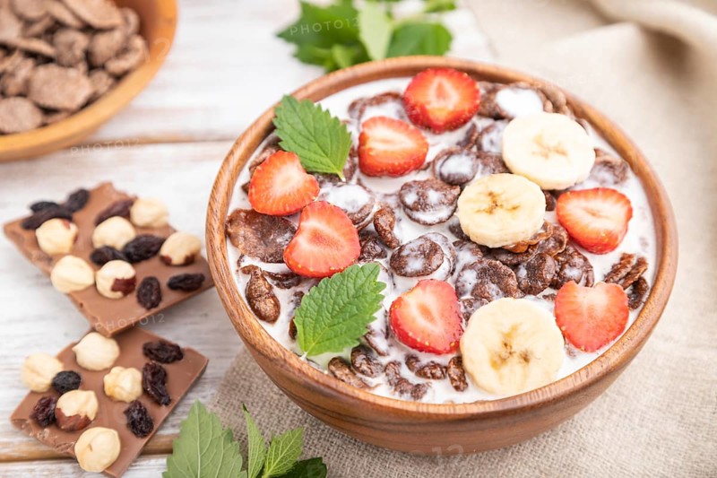 Chocolate cornflakes with milk and strawberry in wooden bowl on white wooden background and linen textile. Side view, close up, selective focus