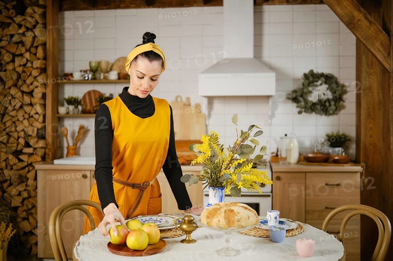 A woman in a yellow sleeveless sundress takes an apple from a tray standing in country style kitchen