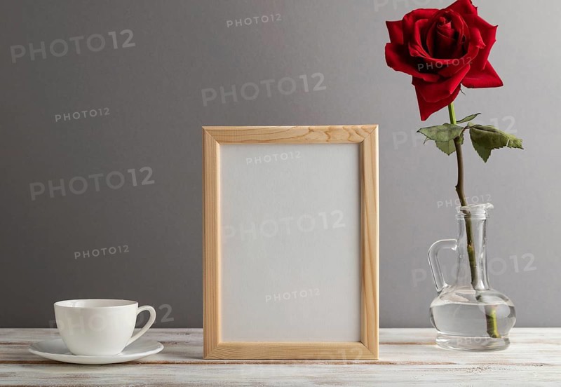 Beige wooden frame mockup with red rose in glass and white coffee cup on gray paper background. Blank, vertical orientation, still life, copy space