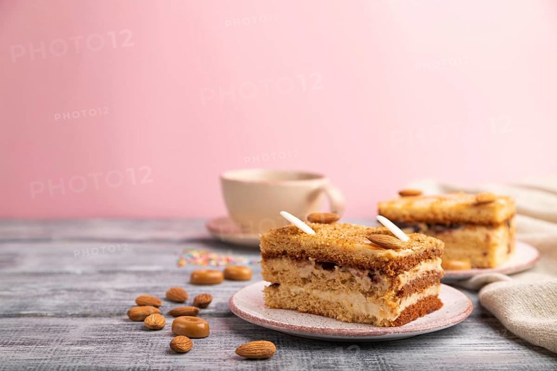 Honey cake with milk cream, caramel, almonds and a cup of coffee on a gray and pink background and linen textile. Side view, close up, selective focus, copy space