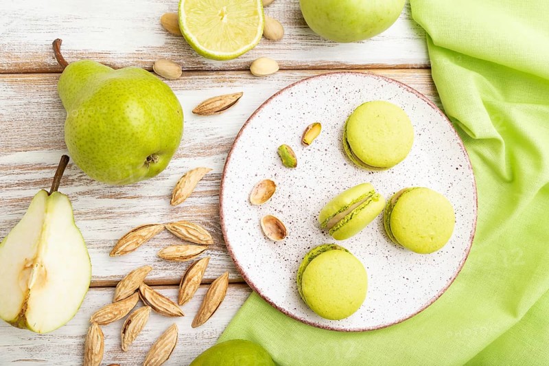 Green macarons or macaroons cakes with cup of coffee on a white wooden background and green linen textile. Top view, flat lay, close up